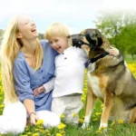 Femme joyeuse avec enfant et chien dans un parc ensoleillé, moment de bonheur familial.