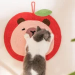 Un chat noir et blanc joue sur un tapis pour chat en forme de pomme rouge.